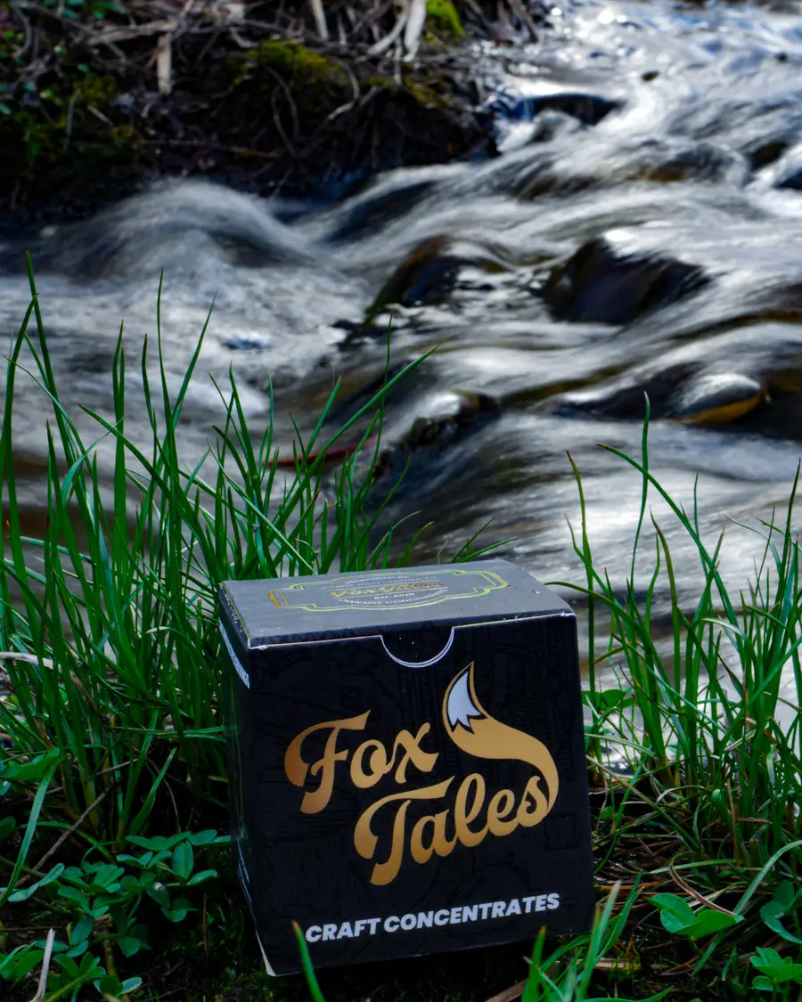 A Fox Tales craft concentrates carton in grass beside a running stream, portrait crop, the moving water blurred behind it.