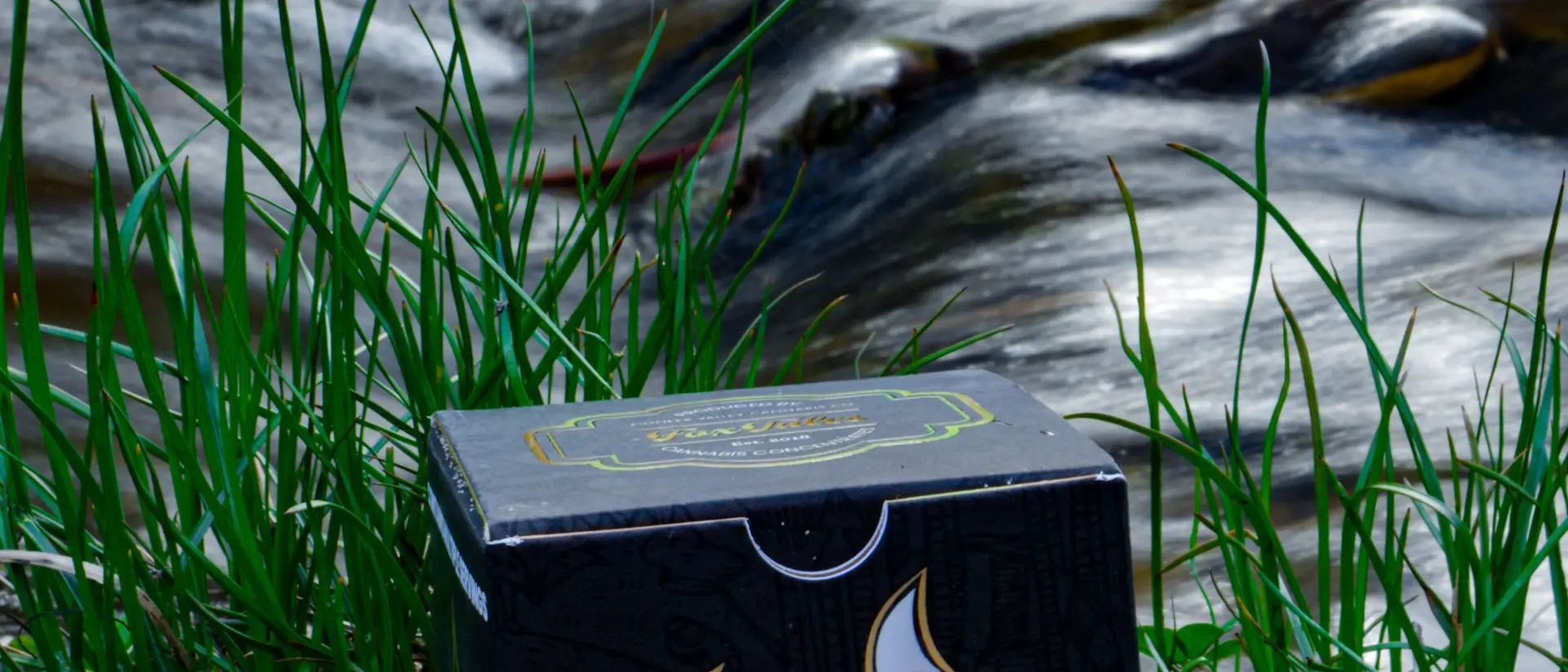 A Fox Tales craft concentrates carton standing in grass on the bank of a fast-running stream, the water blurred to white behind it.