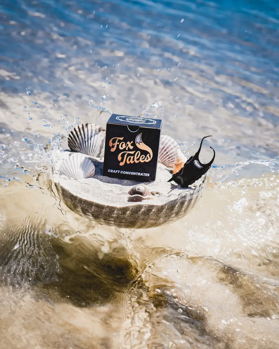 A Fox Tales craft concentrates carton standing in a glass dish of sand in breaking surf, portrait crop, with water splashing around the shells beside it.