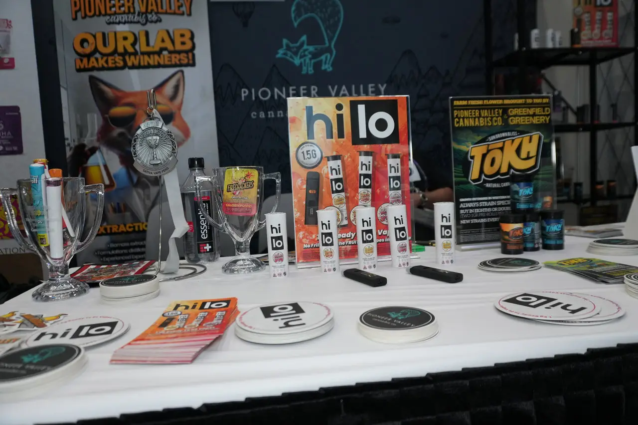 A trade-show table laid out with a Hi Lo counter display card, a Tokh table tent, Pioneer Valley coasters and sell sheets, in front of the Our Lab Makes Winners backdrop banner.