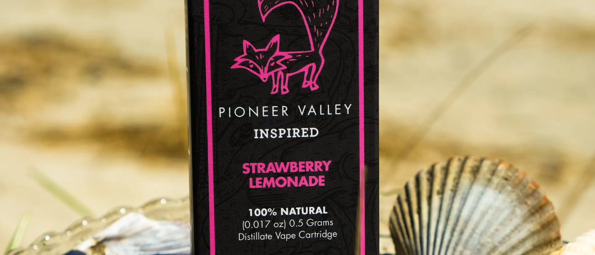 A Pioneer Valley Inspired Strawberry Lemonade vape cartridge carton standing in a glass dish of sand on a beach, scallop shells piled around its base and dune grass out of focus behind it.