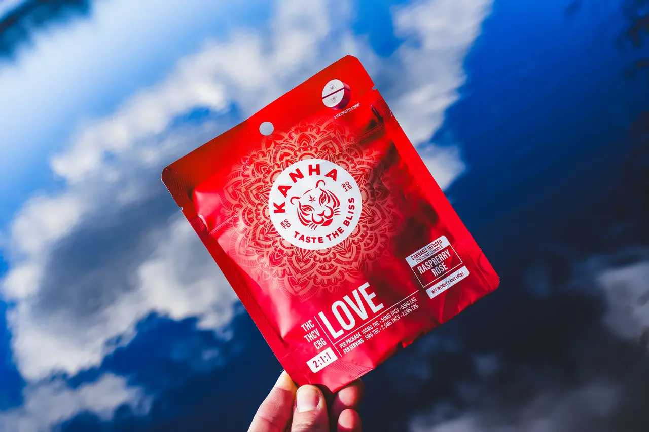 A hand holding a red Kanha Love raspberry rose gummies pouch up against a bright blue sky with broken cloud behind it.